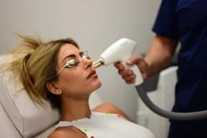 Woman lying in a reclining medical chair wearing protective goggles as a clinician uses a handheld device near her face for a cosmetic procedure